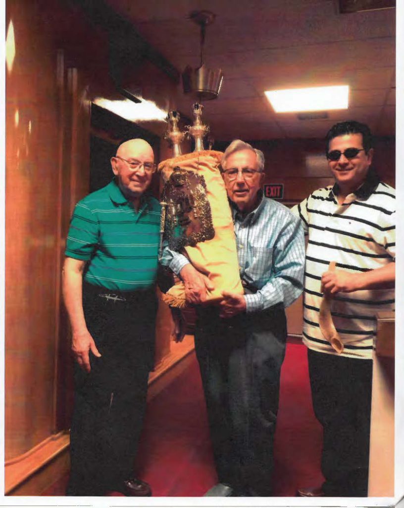 Photograph of three white men smiling. The man in the center is holding a Torah scroll with a yellow Torah cover.